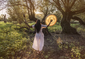 Shaman woman hands wide spread A woman in white dress holding an object near trees.